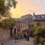 Edinburgh Castle seen at sunset from a steep Old Town street, framed by historic stone buildings, leafy trees and greenery. Two people walk downhill in the foreground beneath a warm orange and pink sky.