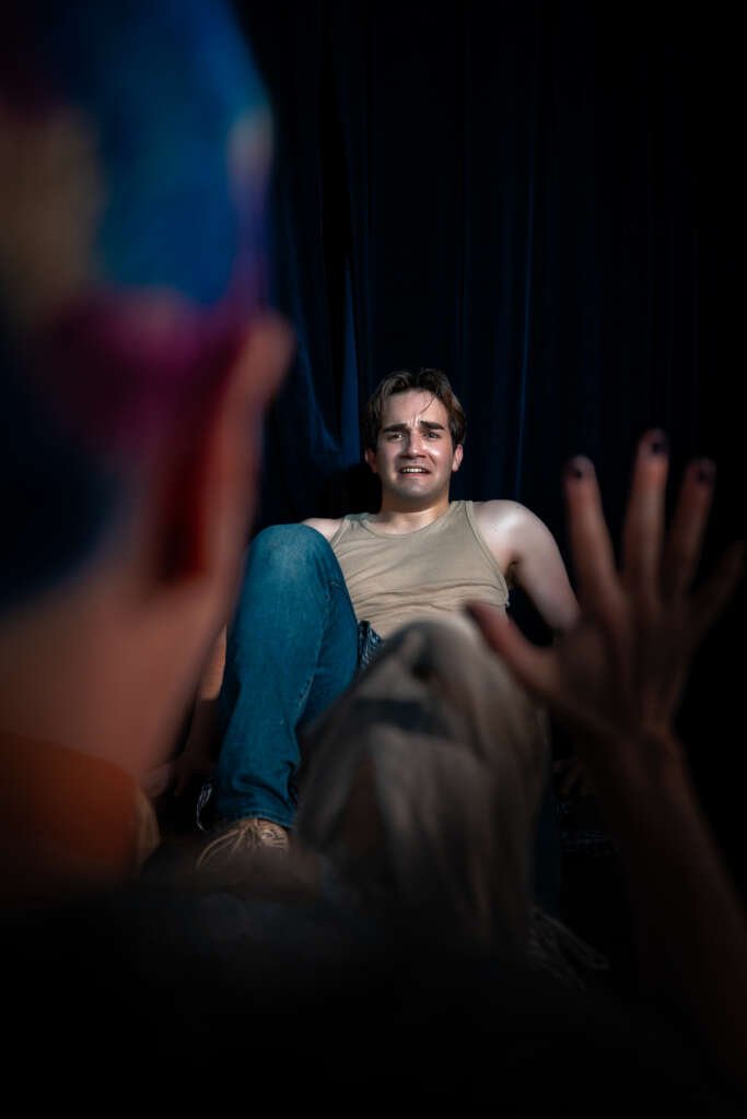 A performer in a beige vest and jeans sits on the floor against dark curtains, looking uneasy towards another person in the blurred foreground who has one hand raised.