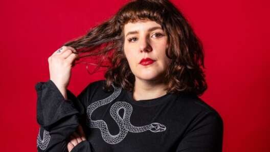 Portrait of a person with wavy brown hair and red lipstick against a bright red background, wearing a black top with a white snake design and holding a strand of their hair.