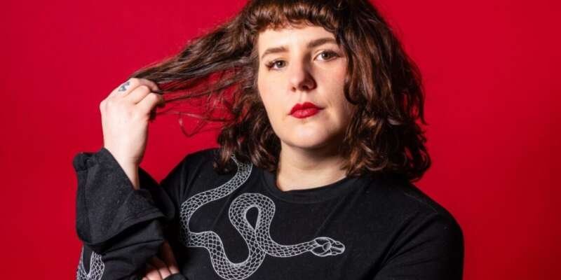 Portrait of a person with wavy brown hair and red lipstick against a bright red background, wearing a black top with a white snake design and holding a strand of their hair.