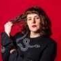Portrait of a person with wavy brown hair and red lipstick against a bright red background, wearing a black top with a white snake design and holding a strand of their hair.
