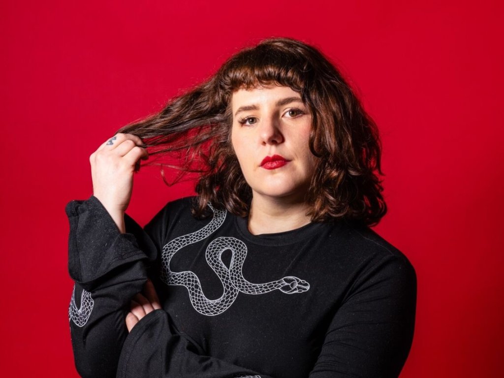 Portrait of a person with wavy brown hair and red lipstick against a bright red background, wearing a black top with a white snake design and holding a strand of their hair.