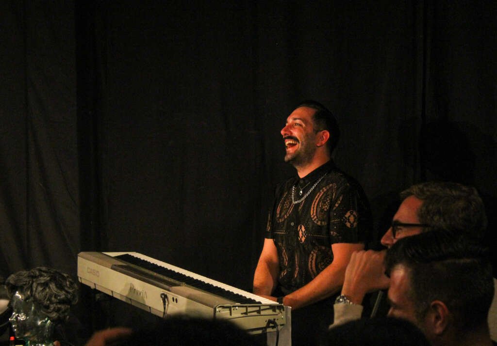 A smiling performer in a black lace top sits behind an electronic keyboard on a dark stage, laughing as audience members watch nearby.