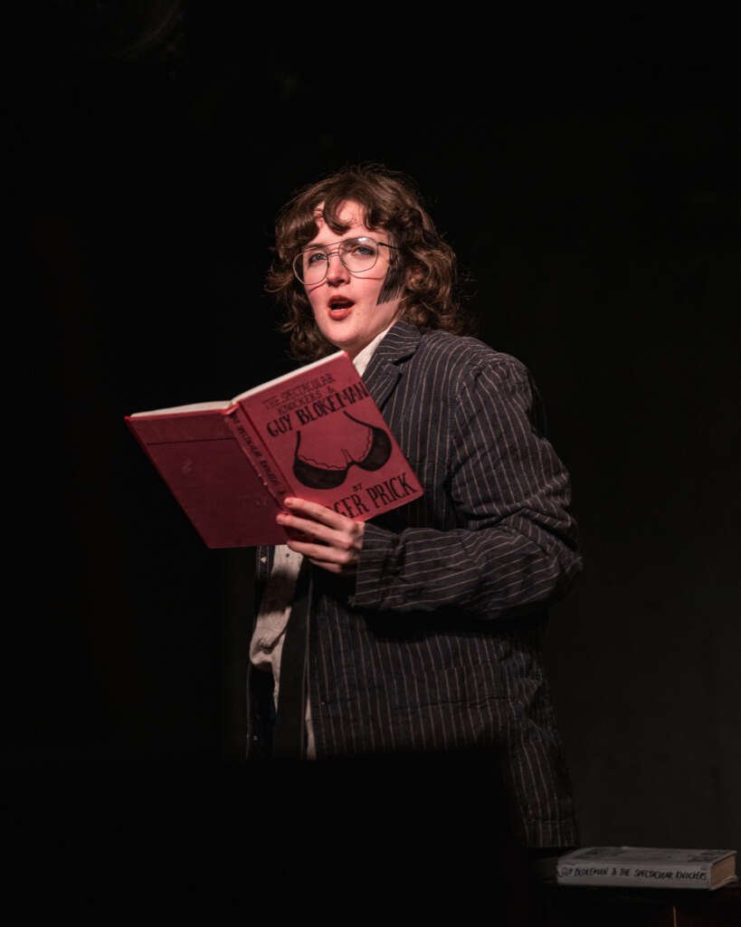 A performer in a pinstripe suit and glasses reads from a red book on a dark stage.