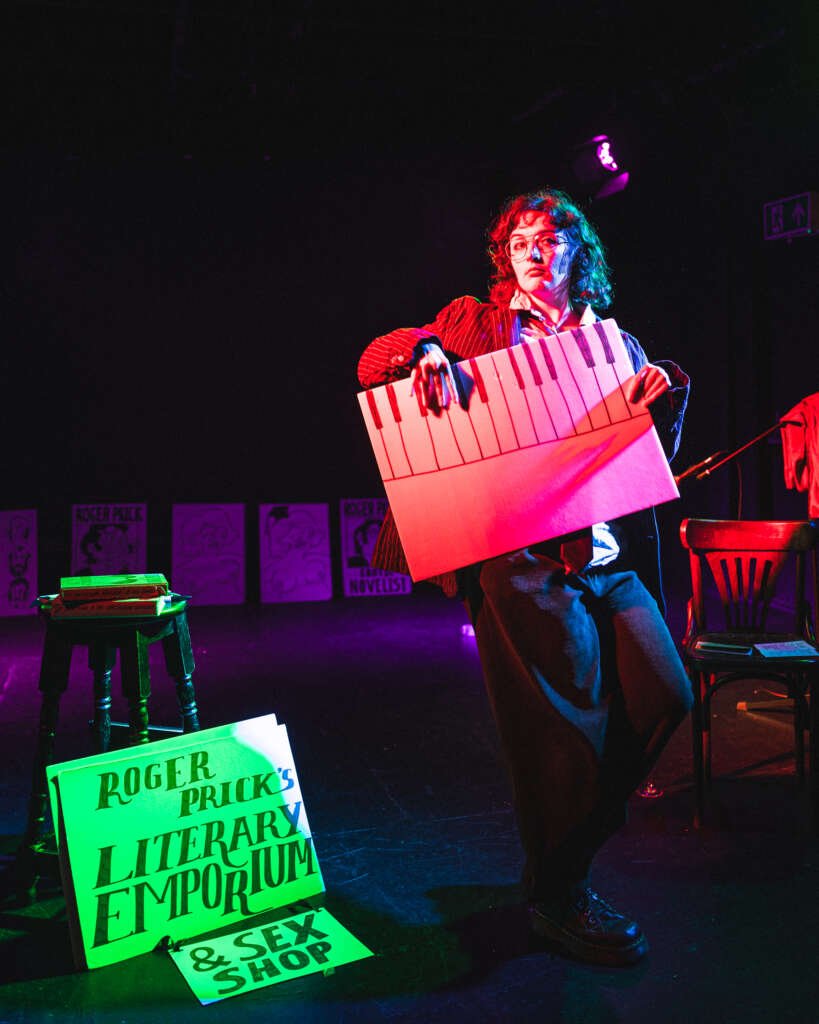 A performer in glasses and a pinstripe suit stands under vivid red, blue and purple stage lighting, holding a large prop designed like piano keys and lips. Signs on the floor read “Roger Prick’s Literary Emporium” and “& Sex Shop.”