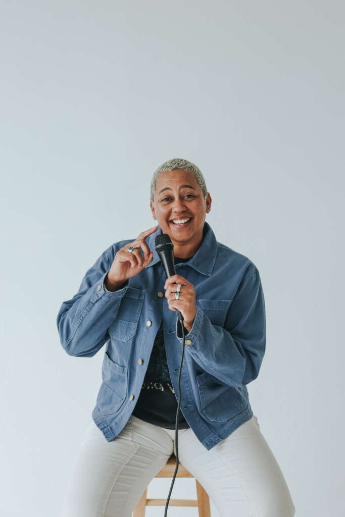 A smiling person with short grey hair sits on a stool, holding a microphone against a pale background.