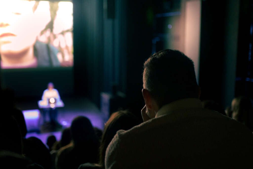 Audience members watch Verona Verbakel perform onstage beneath a large projected video image, seen from the back of the theatre.