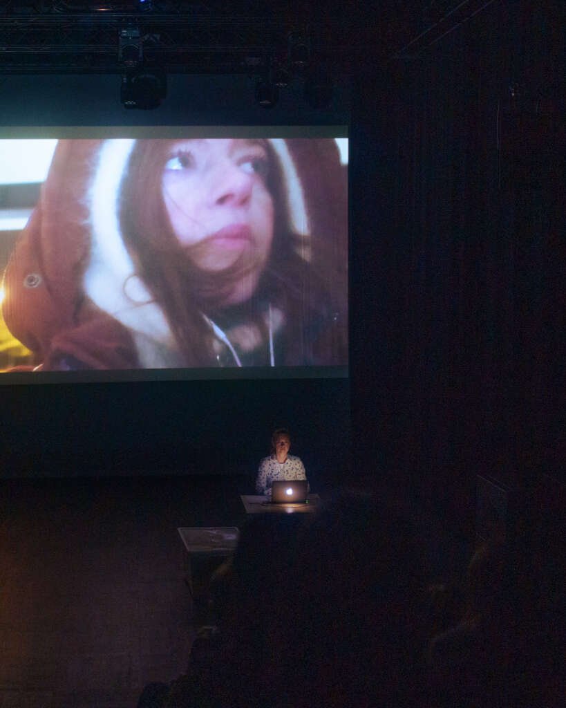 Verona Verbakel performs behind a laptop beneath a large projection showing a close-up video of her face in a hood.