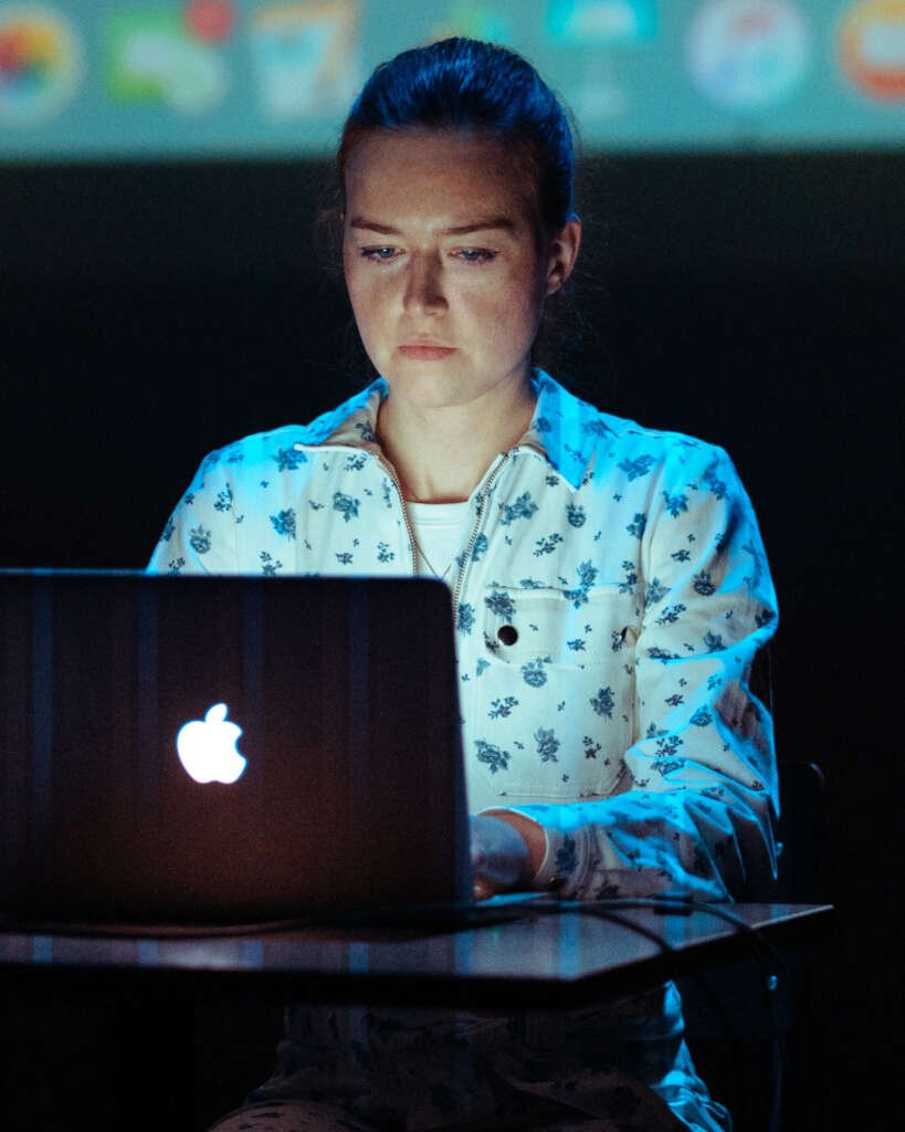 Verona Verbakel sits behind a laptop onstage, looking at the screen under blue lighting, with a projection visible behind her.
