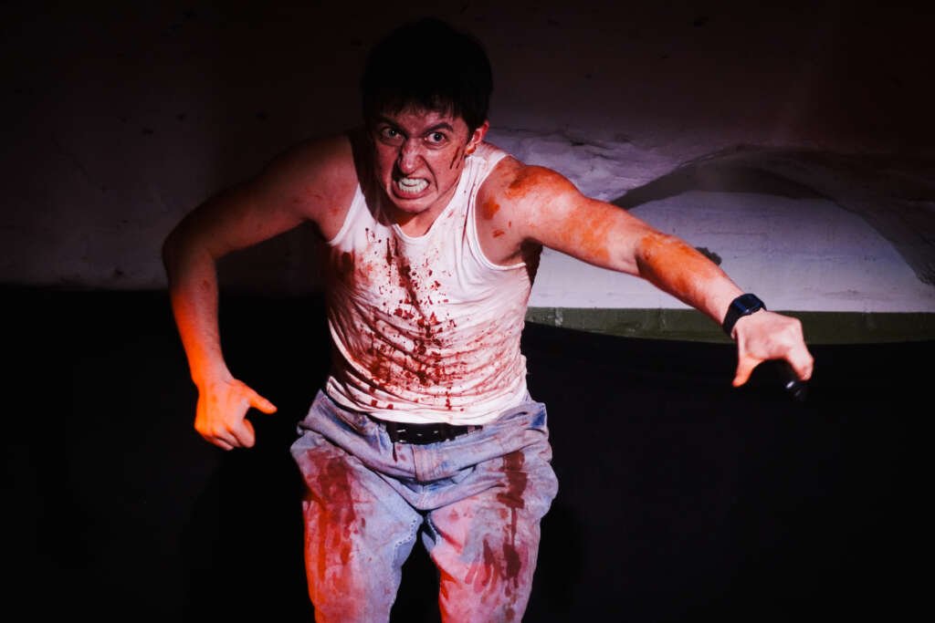 A performer onstage leans forward with an intense expression, wearing a white vest and jeans splattered with theatrical blood and gripping a microphone in one hand.