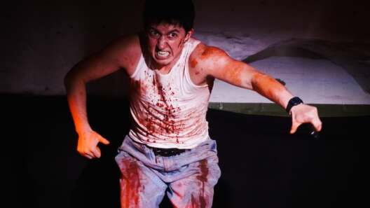 A performer onstage leans forward with an intense expression, wearing a white vest and jeans splattered with theatrical blood and gripping a microphone in one hand.