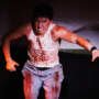 A performer onstage leans forward with an intense expression, wearing a white vest and jeans splattered with theatrical blood and gripping a microphone in one hand.