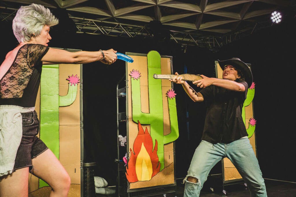 Two performers face off onstage in a comic Wild West-style scene, aiming toy guns at each other in front of cardboard cactus and campfire props.