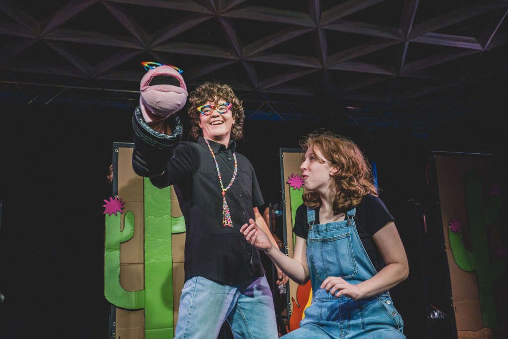 Two performers onstage in front of cardboard cactus scenery. One smiles while holding up a pink hand puppet wearing rainbow glasses, as the other, dressed in denim overalls, looks towards it.