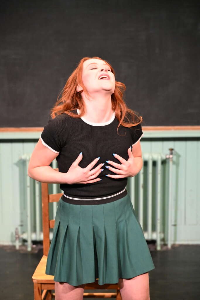 A red-haired woman performs on stage, leaning back with her eyes closed and hands held to her chest, with a wooden chair behind her.