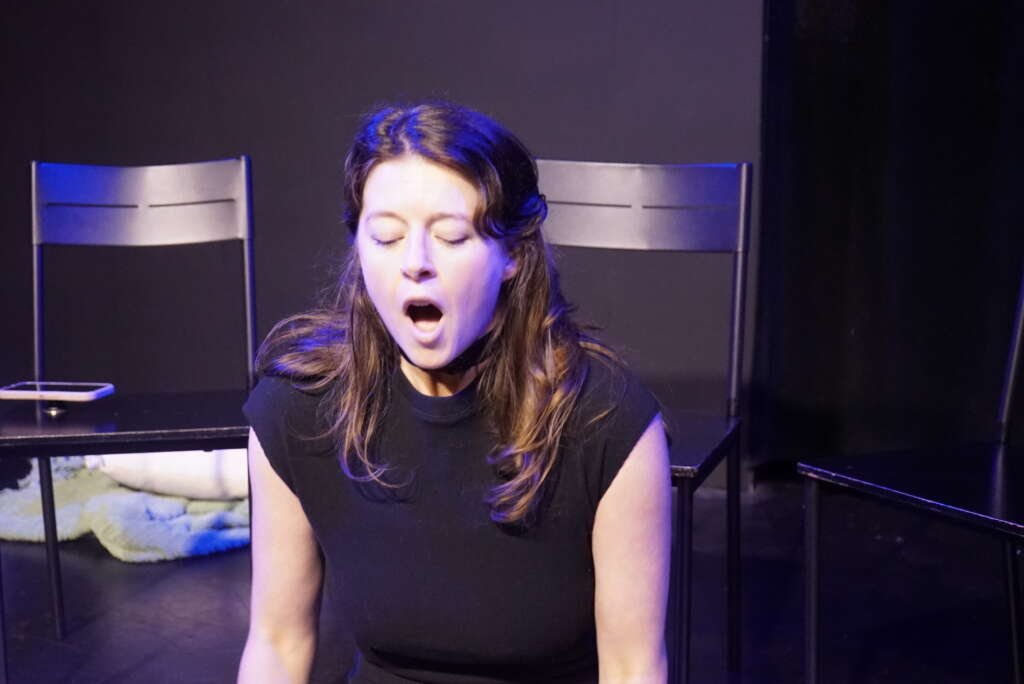 Mimi Collins performs on stage under blue and purple lighting, seated with her eyes closed and mouth open, with several black chairs behind her.