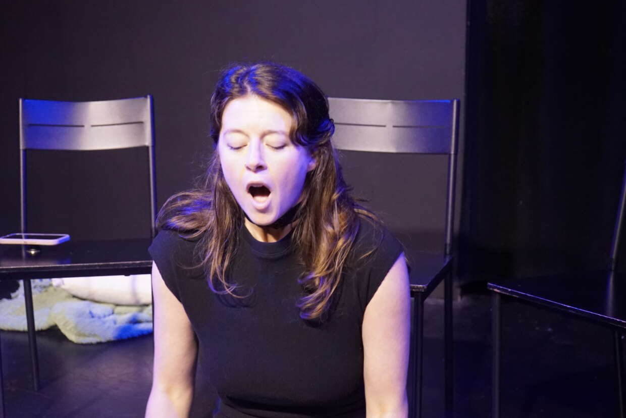 Mimi Collins performs on stage under blue and purple lighting, seated with her eyes closed and mouth open, with several black chairs behind her.