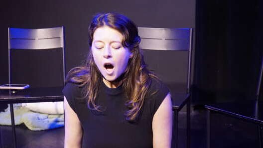 Mimi Collins performs on stage under blue and purple lighting, seated with her eyes closed and mouth open, with several black chairs behind her.