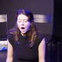 Mimi Collins performs on stage under blue and purple lighting, seated with her eyes closed and mouth open, with several black chairs behind her.