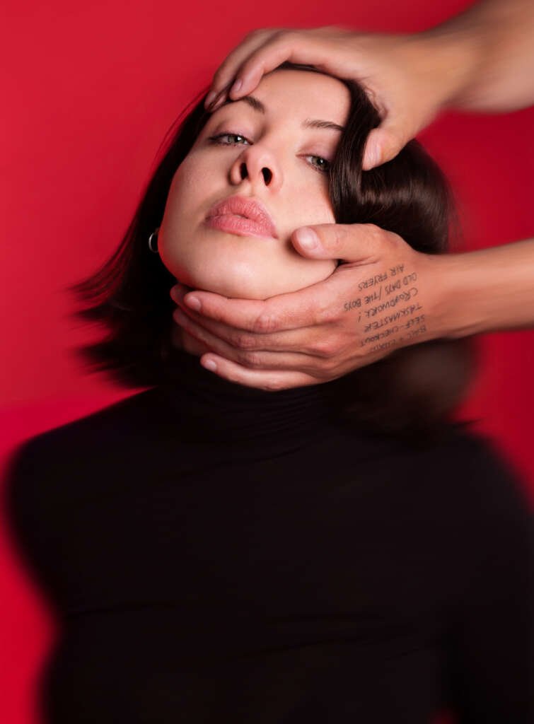 Close-up portrait of a woman against a vivid red background, with two hands framing and holding her face.