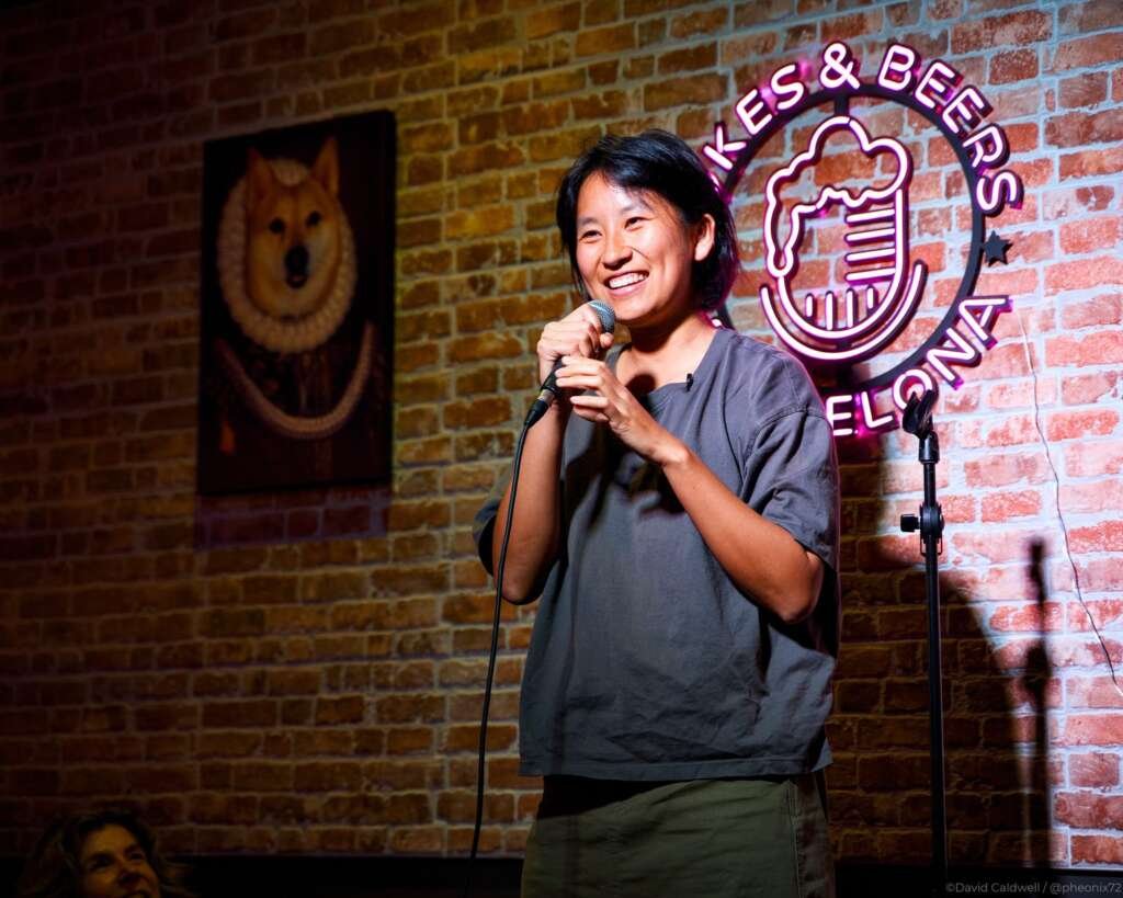 A smiling comedian stands onstage holding a microphone in front of a brick wall, with a glowing “Jokes & Beers” neon sign behind them.