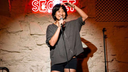 A smiling comedian stands onstage holding a microphone, one hand raised above their head, in front of a stone wall and a glowing red “SECRET” sign.