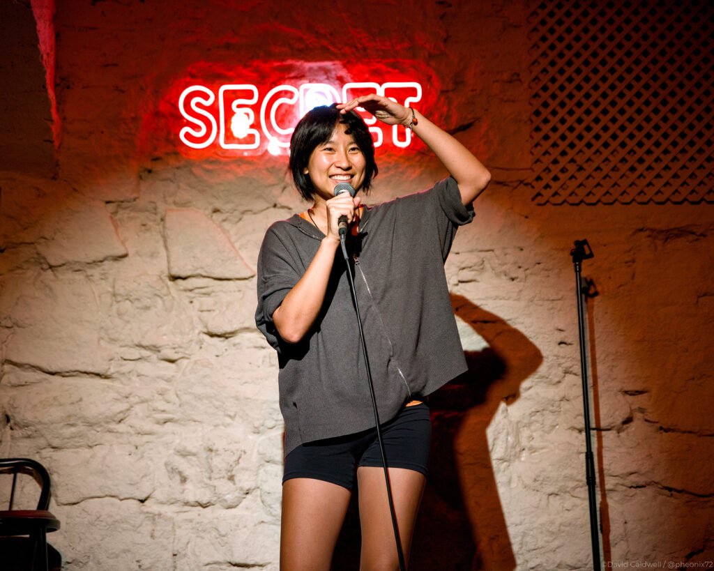 A smiling comedian stands onstage holding a microphone, one hand raised above their head, in front of a stone wall and a glowing red “SECRET” sign.