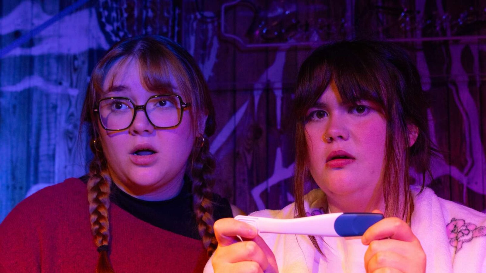 Two performers stare ahead with surprised expressions under purple and blue stage lighting. One wears glasses and has braided hair, while the other holds a pregnancy test in both hands.