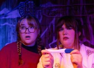 Two performers stare ahead with surprised expressions under purple and blue stage lighting. One wears glasses and has braided hair, while the other holds a pregnancy test in both hands.