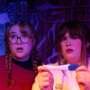 Two performers stare ahead with surprised expressions under purple and blue stage lighting. One wears glasses and has braided hair, while the other holds a pregnancy test in both hands.