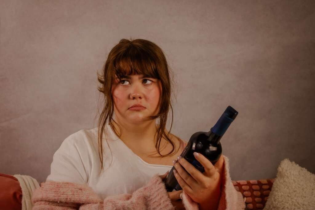 performer sits on a sofa holding a bottle of wine, looking off to the side with an uneasy expression while wrapped in a pink cardigan.