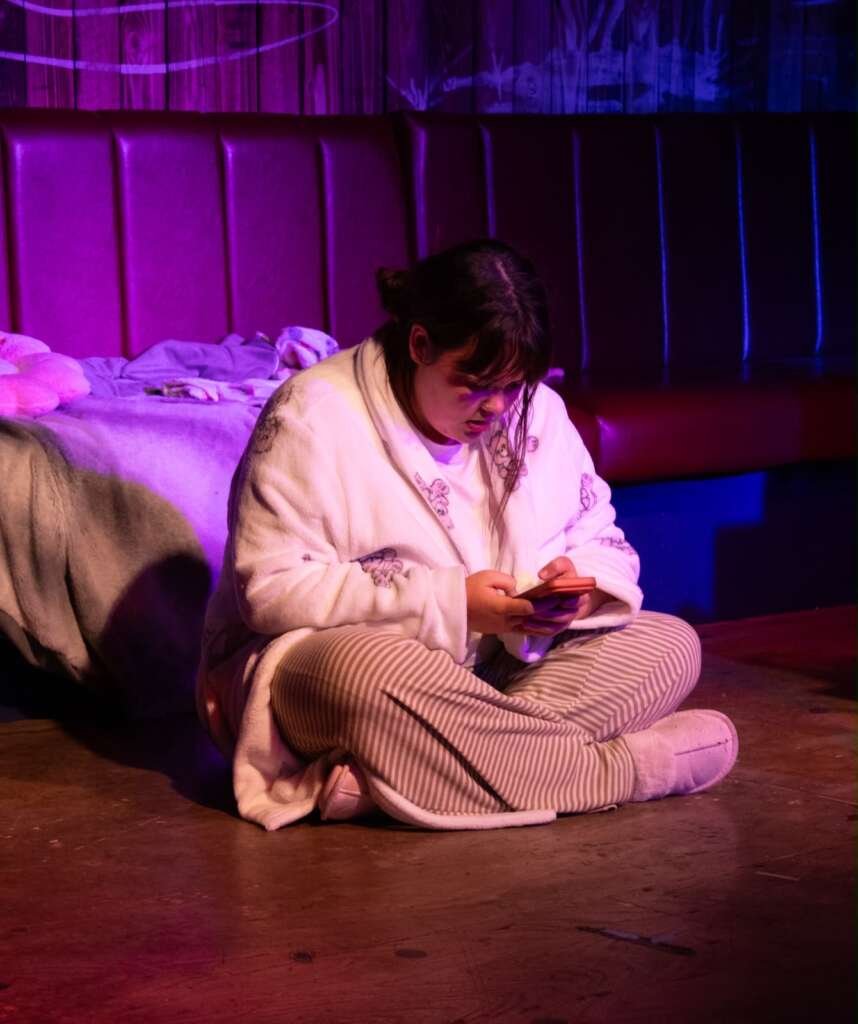A performer in a white dressing gown and striped pyjamas sits cross-legged on the floor beside a bed, looking down at a phone under purple and blue stage lighting.
