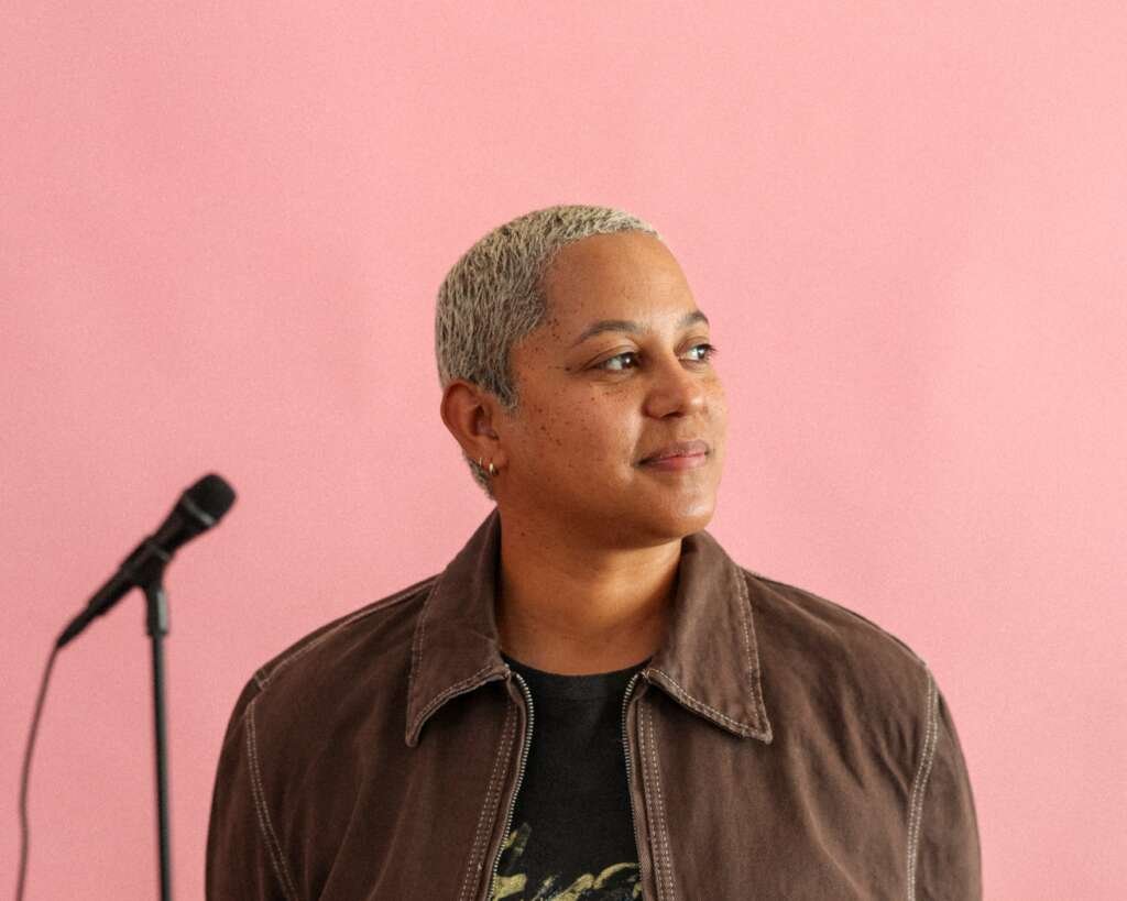 Portrait of a person with closely cropped grey hair wearing a brown jacket, standing beside a microphone against a plain pink background.