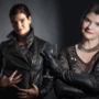 Melissa Todd appears twice in a composite studio portrait against a dark background. On the left, she wears a black leather jacket and gloves and holds a wine glass; on the right, she wears a black patterned top with a chunky silver necklace. In both portraits, she looks towards the camera.