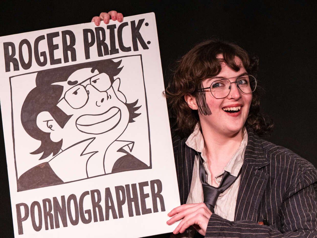 A smiling performer in glasses, a pinstripe jacket and loose tie holds a large illustrated sign reading “Roger Prick: Pornographer” against a black background.