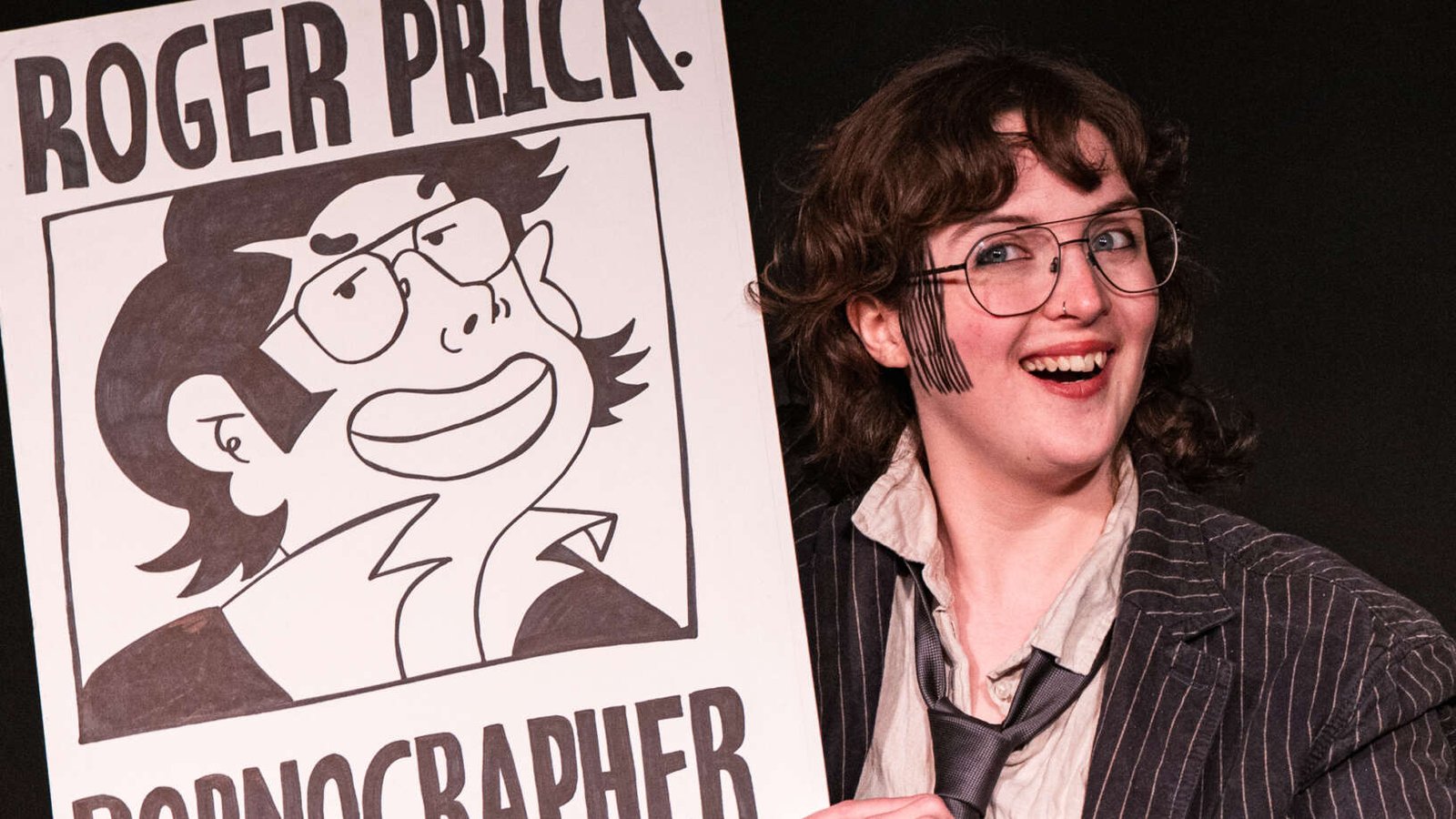 A smiling performer in glasses, a pinstripe jacket and loose tie holds a large illustrated sign reading “Roger Prick: Pornographer” against a black background.