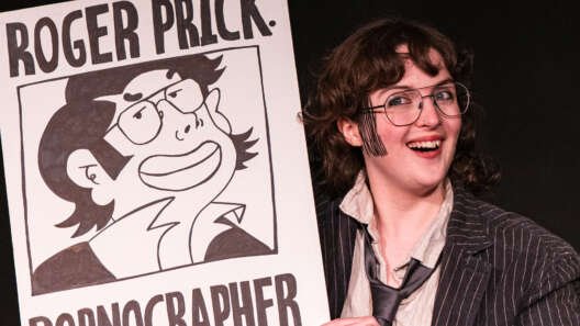 A smiling performer in glasses, a pinstripe jacket and loose tie holds a large illustrated sign reading “Roger Prick: Pornographer” against a black background.