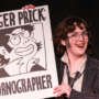 A smiling performer in glasses, a pinstripe jacket and loose tie holds a large illustrated sign reading “Roger Prick: Pornographer” against a black background.