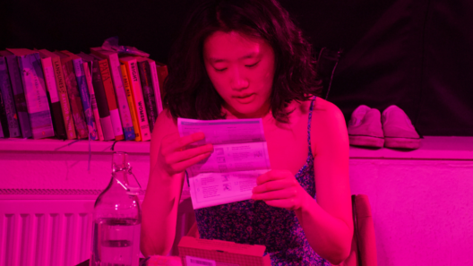 A performer sits at a table under vivid pink stage lighting, reading an information leaflet beside a glass water bottle and a medical testing kit, with books and shoes in the background.
