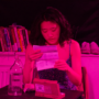 A performer sits at a table under vivid pink stage lighting, reading an information leaflet beside a glass water bottle and a medical testing kit, with books and shoes in the background.