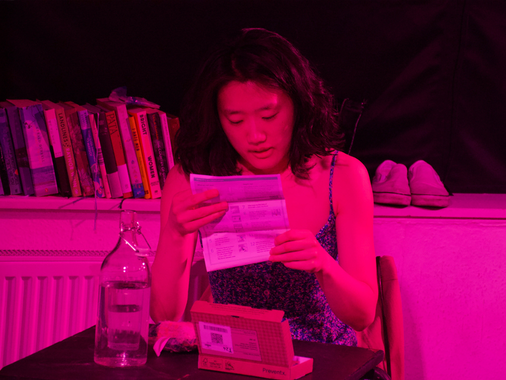 A performer sits at a table under vivid pink stage lighting, reading an information leaflet beside a glass water bottle and a medical testing kit, with books and shoes in the background.