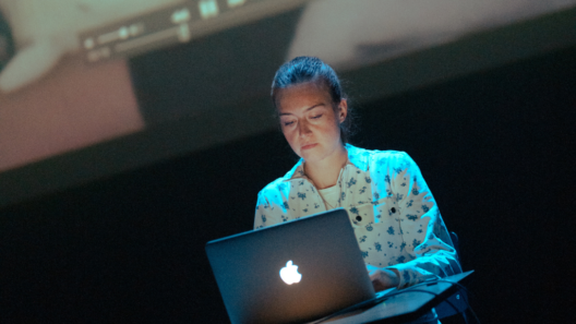 Verona Verbakel sits behind a laptop on a dark stage, lit in blue, with a projected video playing above her.
