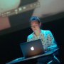 Verona Verbakel sits behind a laptop on a dark stage, lit in blue, with a projected video playing above her.