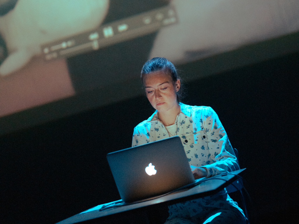 Verona Verbakel sits behind a laptop on a dark stage, lit in blue, with a projected video playing above her.
