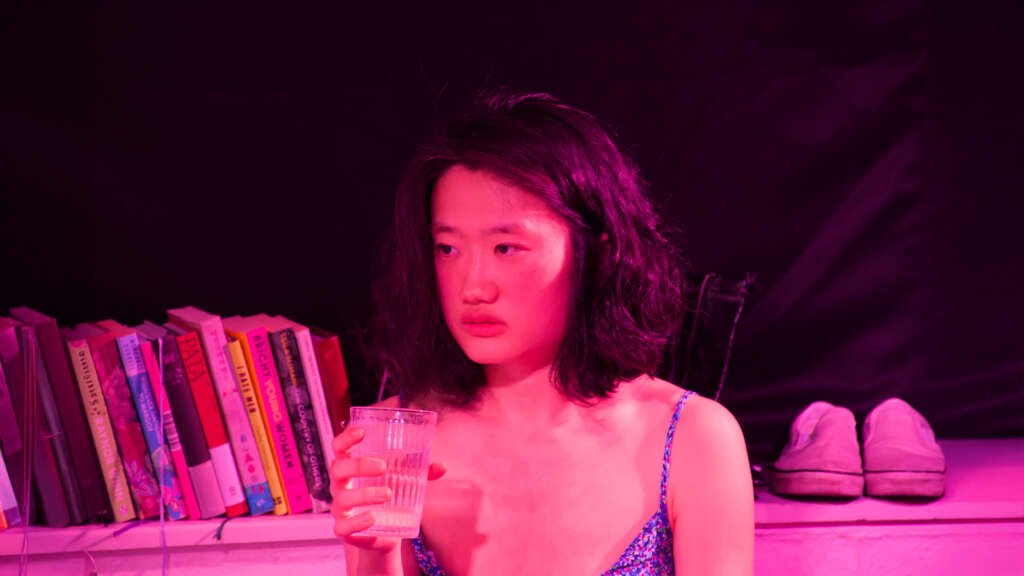A performer under pink stage lighting holds a glass of water, with a row of books and a pair of shoes behind her.