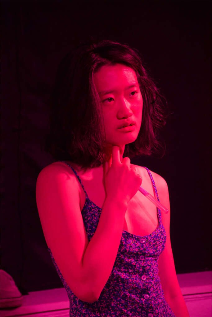A performer under vivid pink stage lighting holds a clear plastic tube against her neck, looking off to one side.