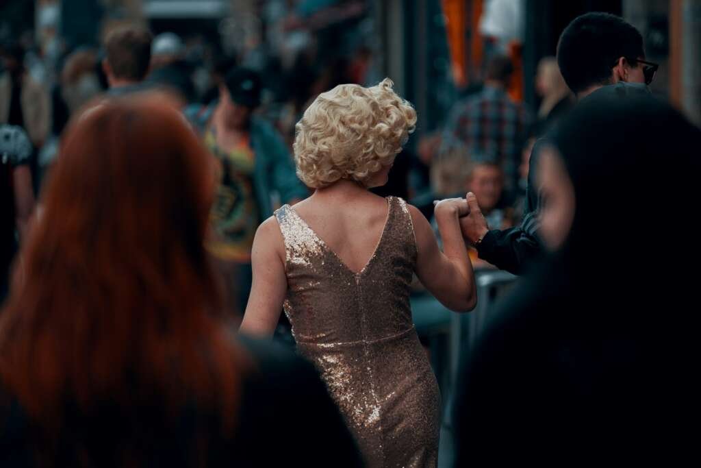 A performer in a sparkling gold dress moves through a crowded street, seen from behind while holding someone’s hand.