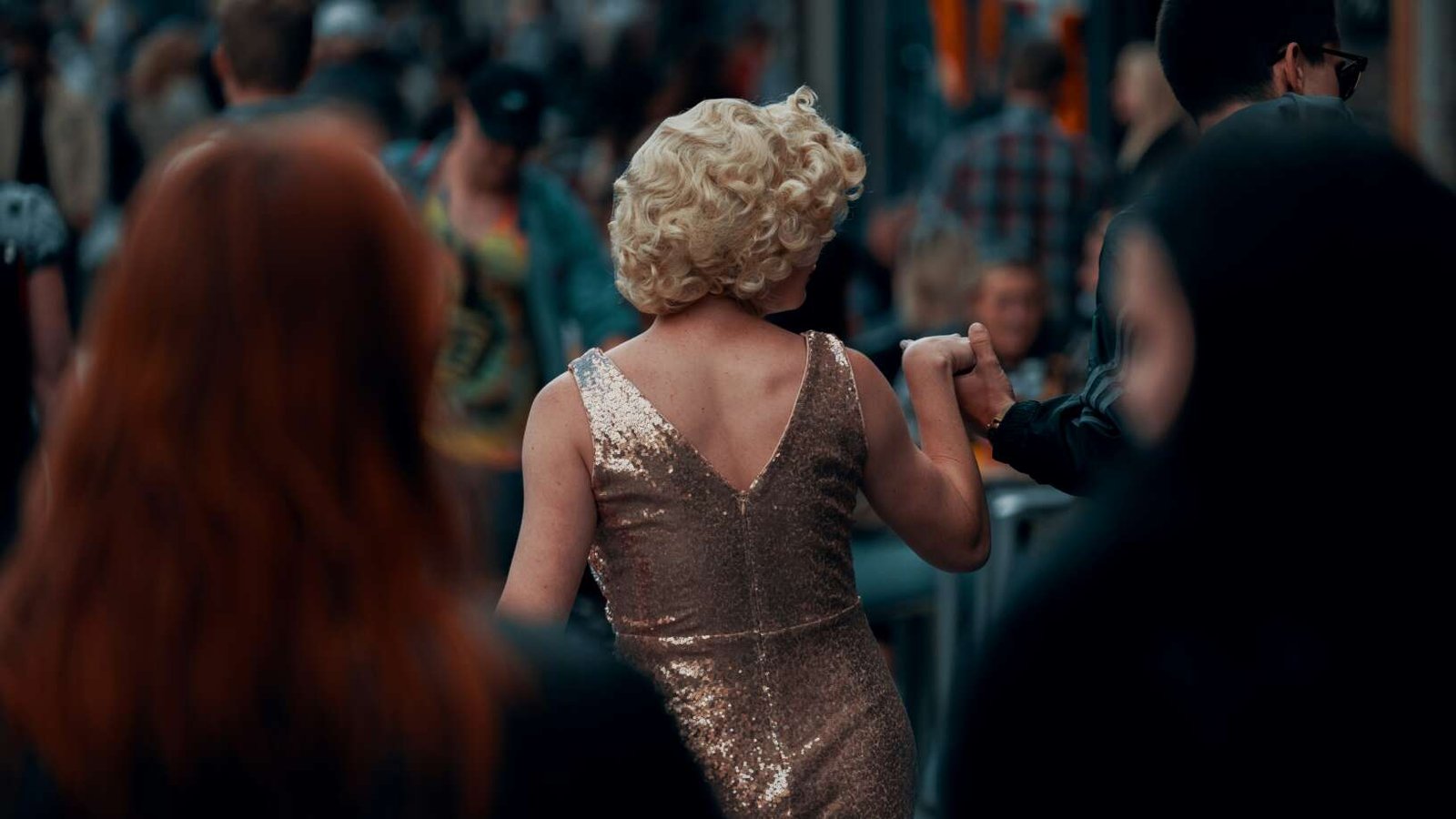 A performer in a sparkling gold dress moves through a crowded street, seen from behind while holding someone’s hand.