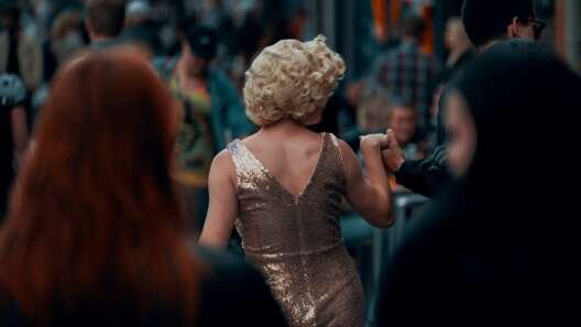 A performer in a sparkling gold dress moves through a crowded street, seen from behind while holding someone’s hand.