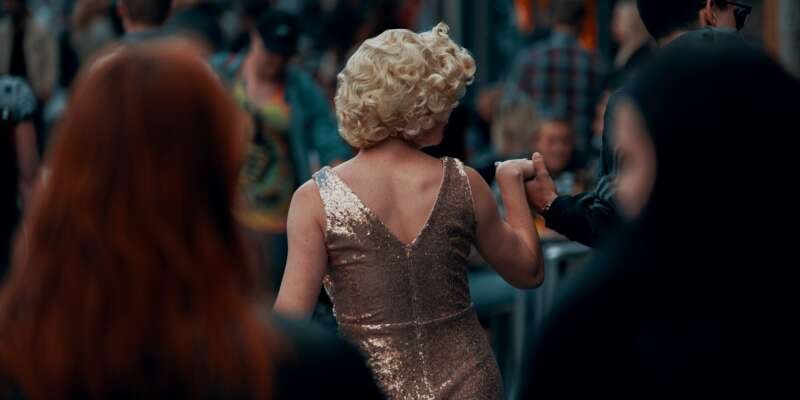 A performer in a sparkling gold dress moves through a crowded street, seen from behind while holding someone’s hand.